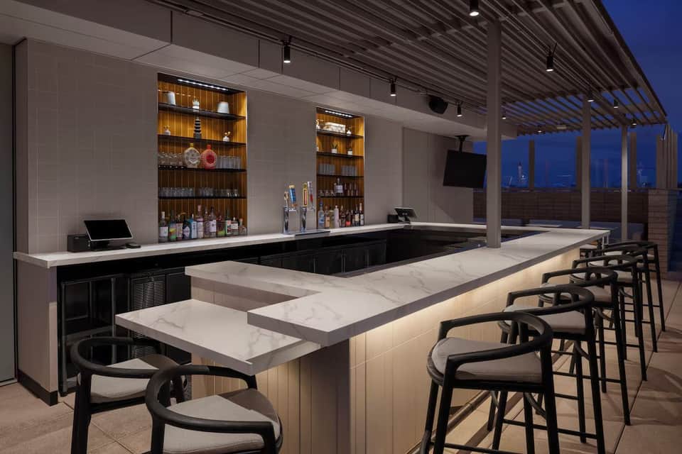 Upscale rooftop bar with marble counters, backlit shelving, and panoramic city views at dusk