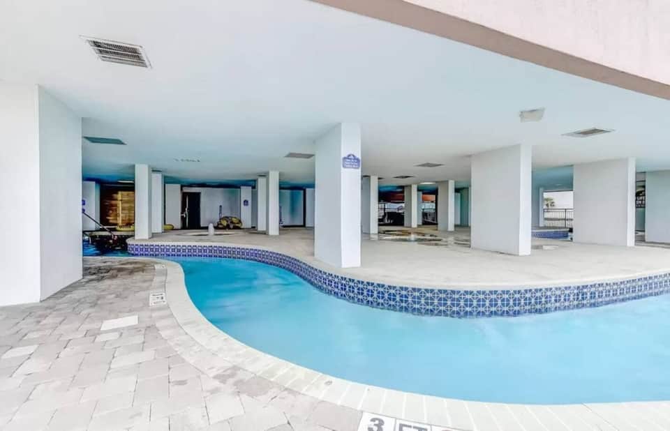 Indoor swimming pool with blue water and decorative tile border, white columns, and spacious facility with multiple entrances