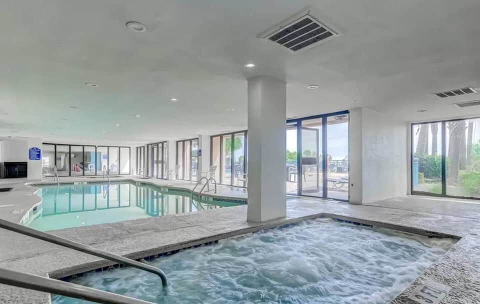 Indoor pool area with hot tub in foreground, large windows overlooking outdoor patio and landscaping, modern resort amenity space