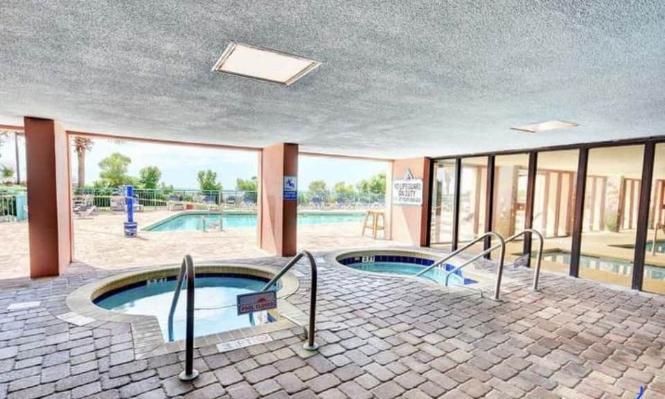 Indoor spa area with two hot tubs, columns, large windows, outdoor pool visible, and glass patio doors to beach or grounds