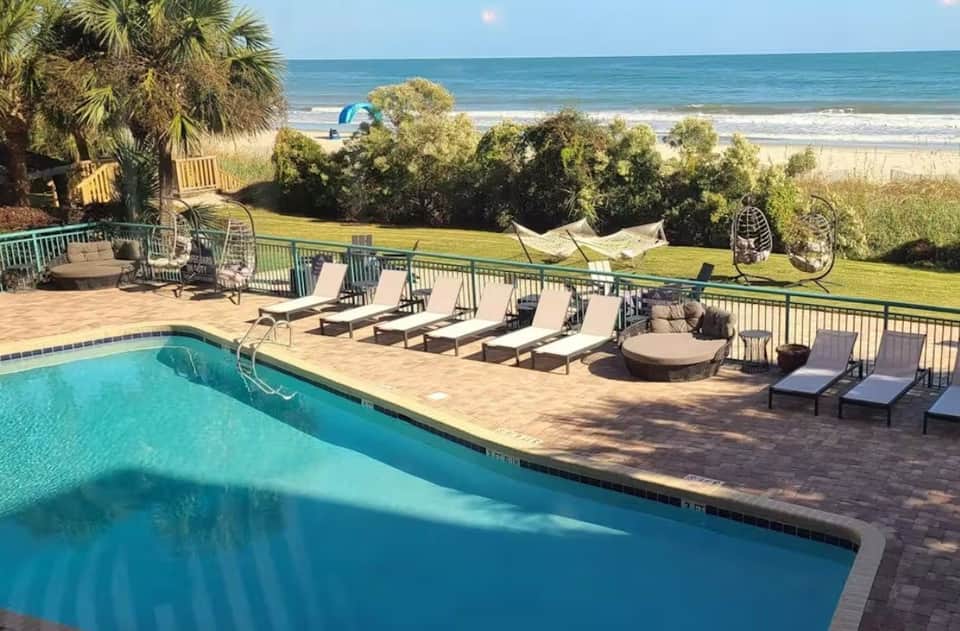 Oceanfront pool deck with lounge chairs, palm trees, beach view, and coastal landscape