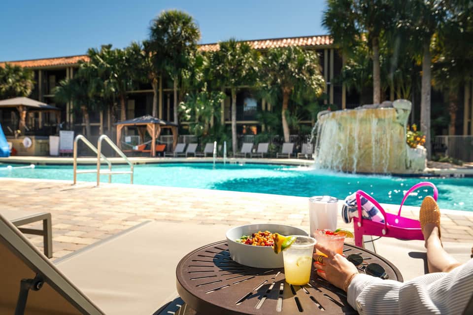 Resort pool with waterfall feature, lounge seating, palm trees, and building with terracotta roof