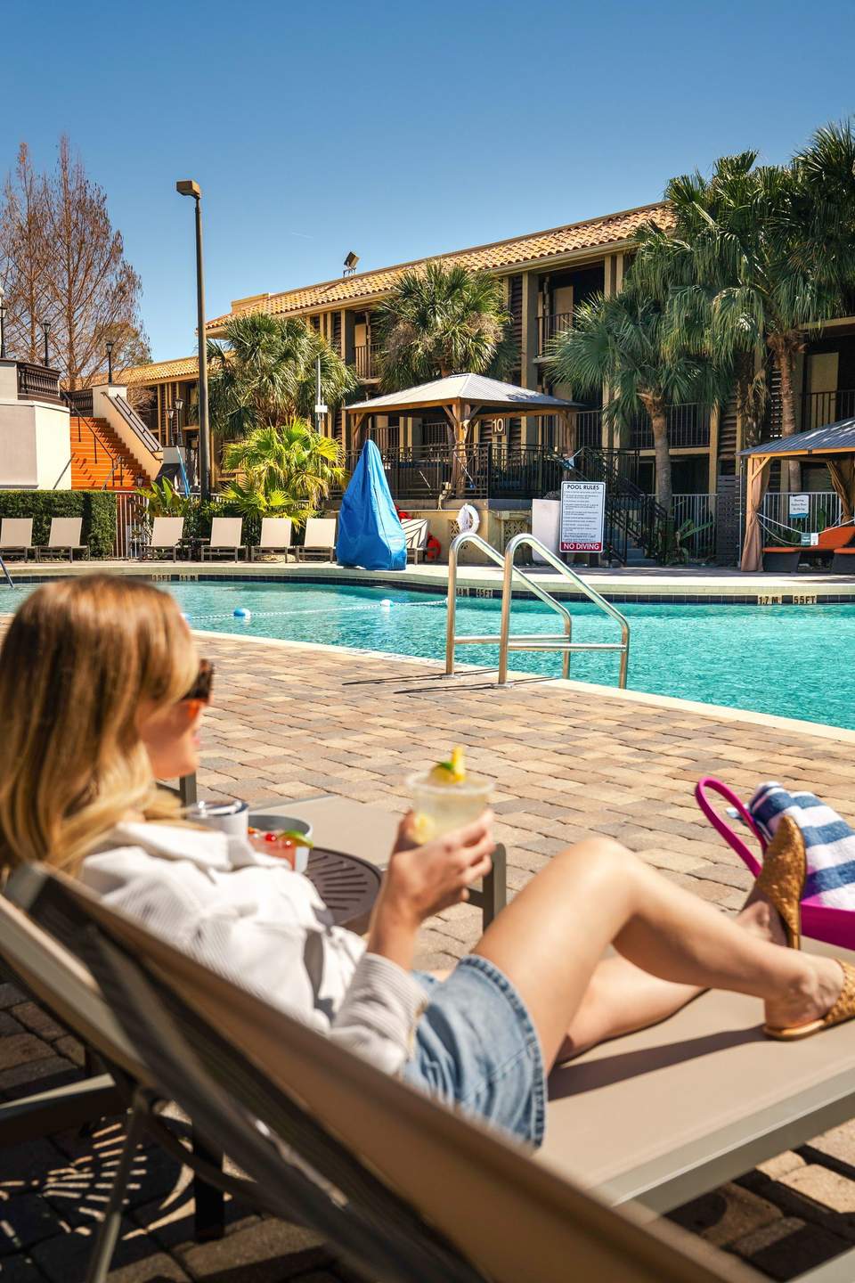 Woman relaxing by resort pool with drink, palm trees and building in background