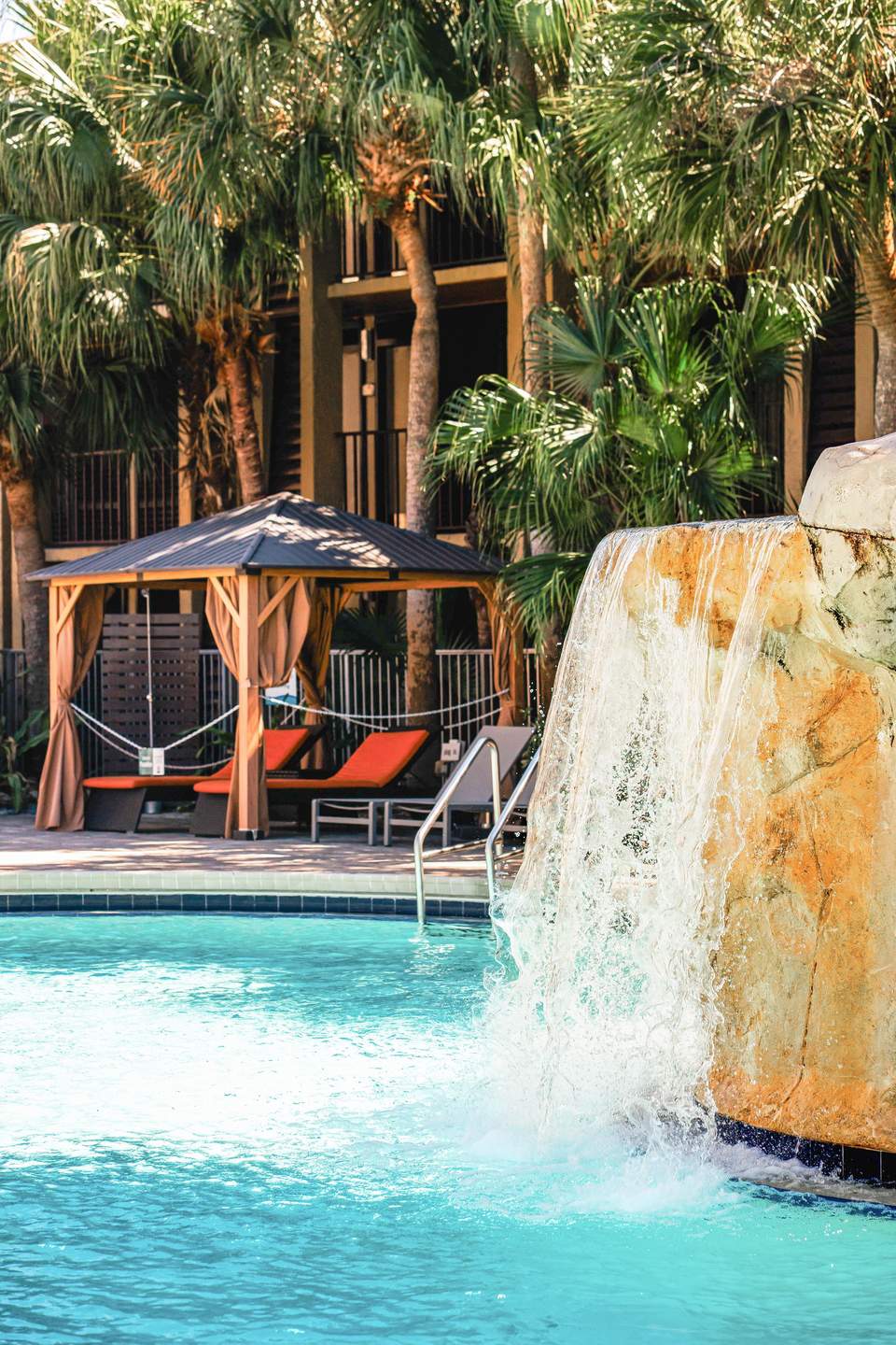 Cascading waterfall feature and wooden cabana with lounge chairs by turquoise pool