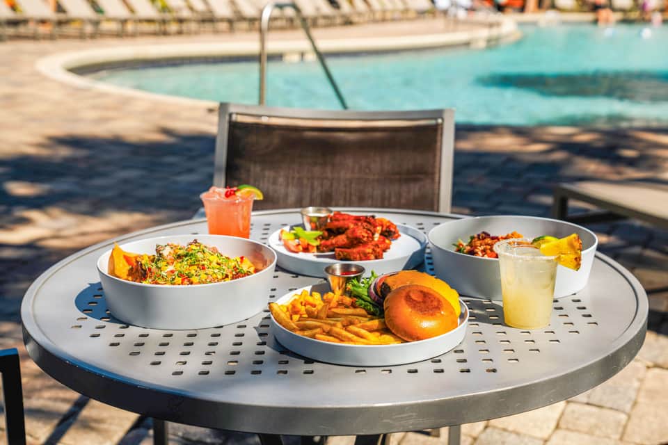 Multiple white bowls of food and drinks arranged on round poolside table with umbrellas