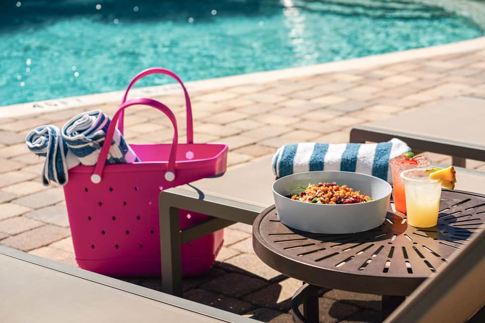 Colorful beach bag and striped towel by poolside table with salad and tropical drinks