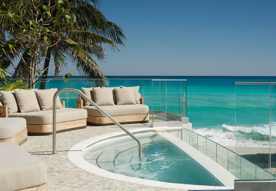 Beachfront infinity pool overlooking turquoise ocean with cream loungers, palm trees, and glass railing on pristine sand