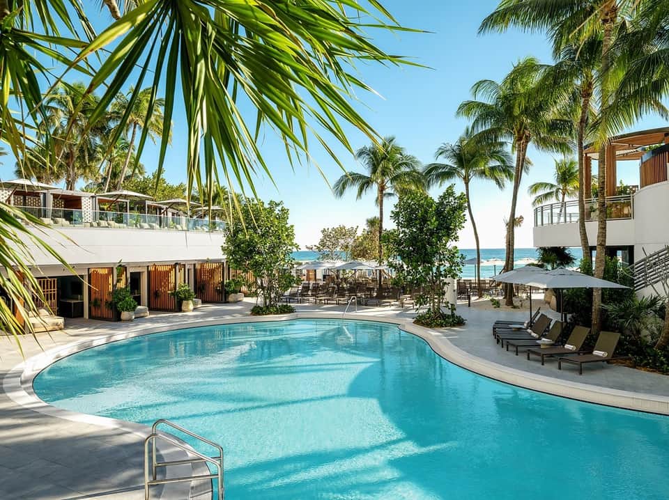 Curved resort pool with palm trees, ocean view, lounge chairs, and beachfront buildings under clear sky