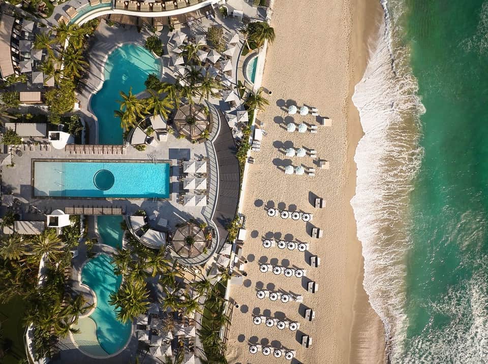 Aerial view of beachfront resort with pools, lounge chairs, palm trees, sand, and turquoise ocean