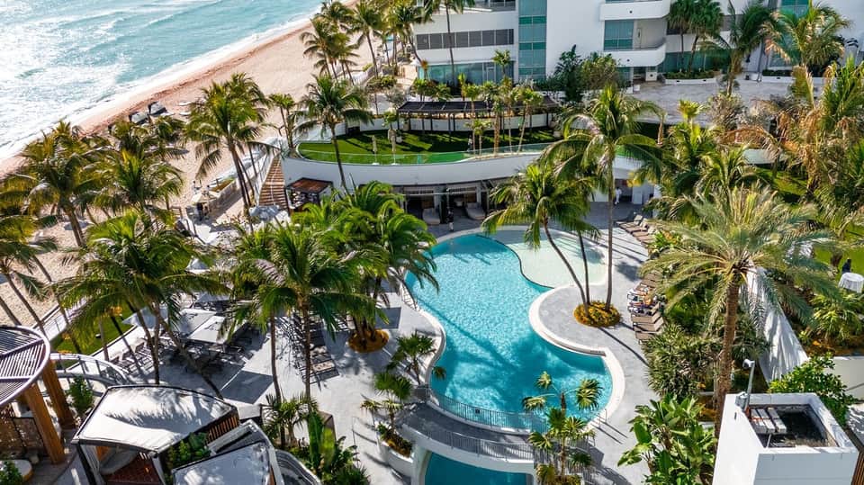 Aerial resort view with curved pool, palm trees, beachfront access, sandy shore, and ocean waves