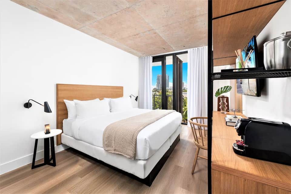 Modern hotel bedroom with wooden headboard, white bedding, concrete ceiling, and balcony overlooking city skyline