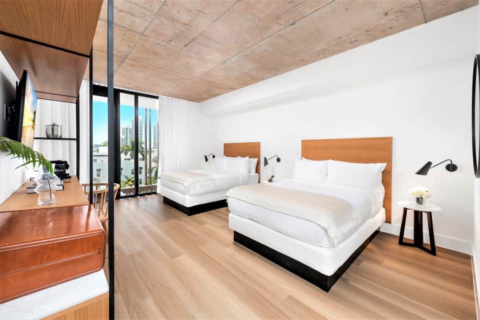 Contemporary hotel suite with two queen beds, wooden headboards, white linens, and floor-to-ceiling windows