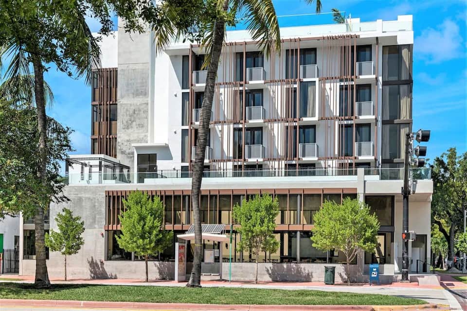 Contemporary multi-story hotel building with white and brown striped facade, ground-level retail, and palm trees
