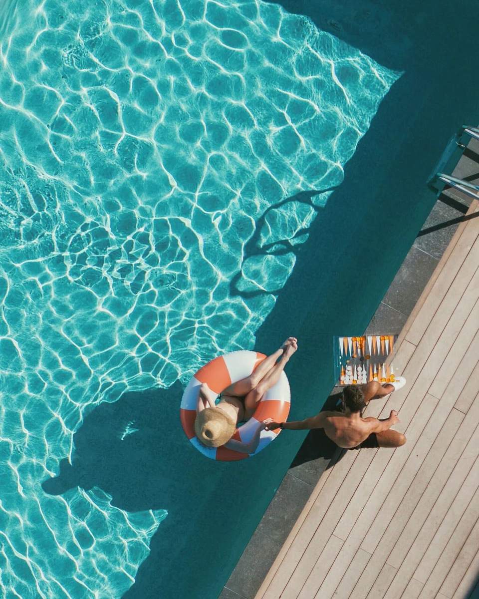 Overhead view of man on orange and white float in turquoise pool next to wooden deck