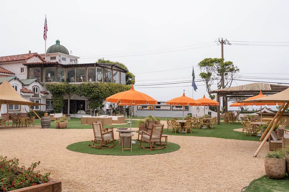 Daytime resort courtyard with orange umbrellas, rocking chairs, fire pit, white building with dome, and grounds
