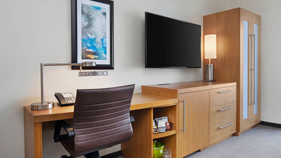 Hotel room workspace with wooden desk, computer monitor, desk chair, and wardrobe cabinet