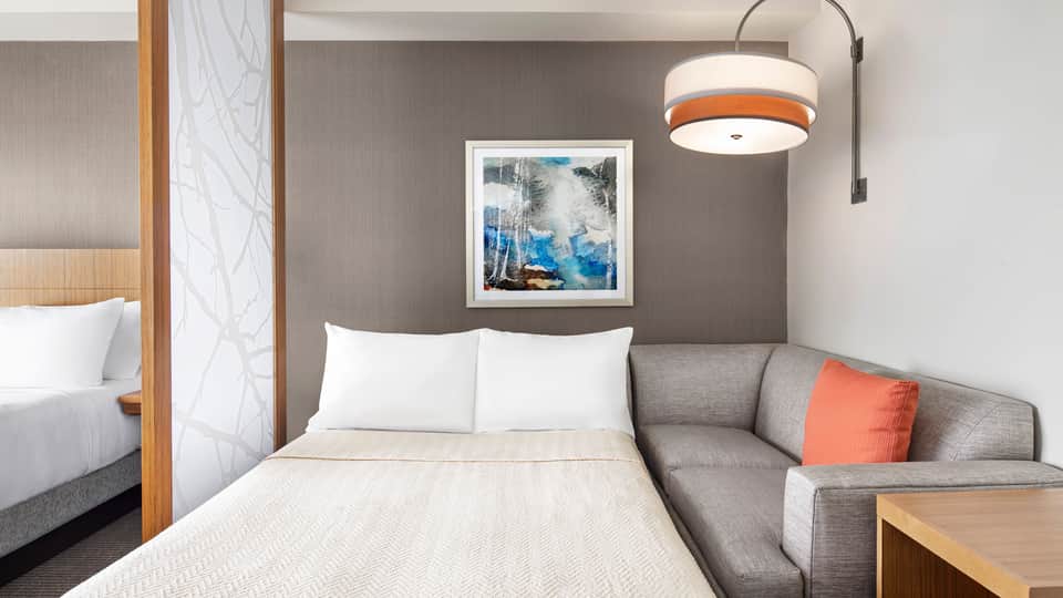 Hotel bedroom with gray accent wall, queen bed, sectional sofa with orange pillow, and pendant lighting