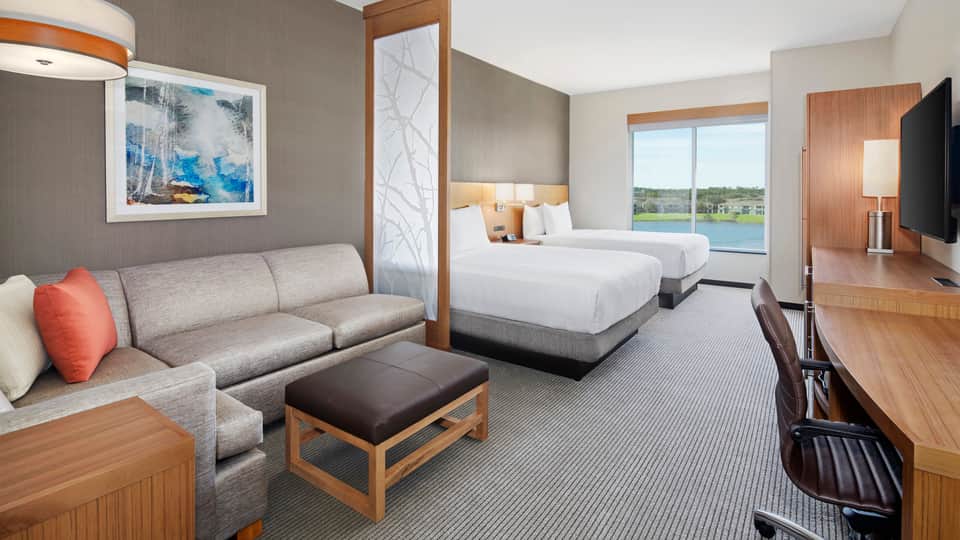 Spacious hotel suite with two queen beds, gray sofa, wood furnishings, large windows overlooking golf course
