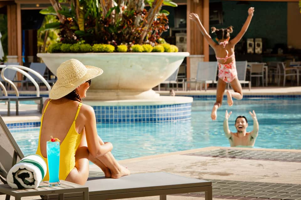 Woman in yellow swimsuit and straw hat relaxing poolside while children play in resort pool