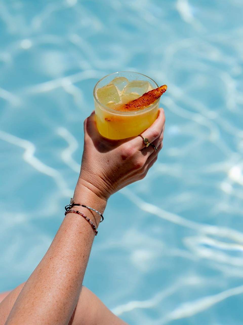Cocktail with citrus garnish held above sparkling blue pool water in bright sunlight