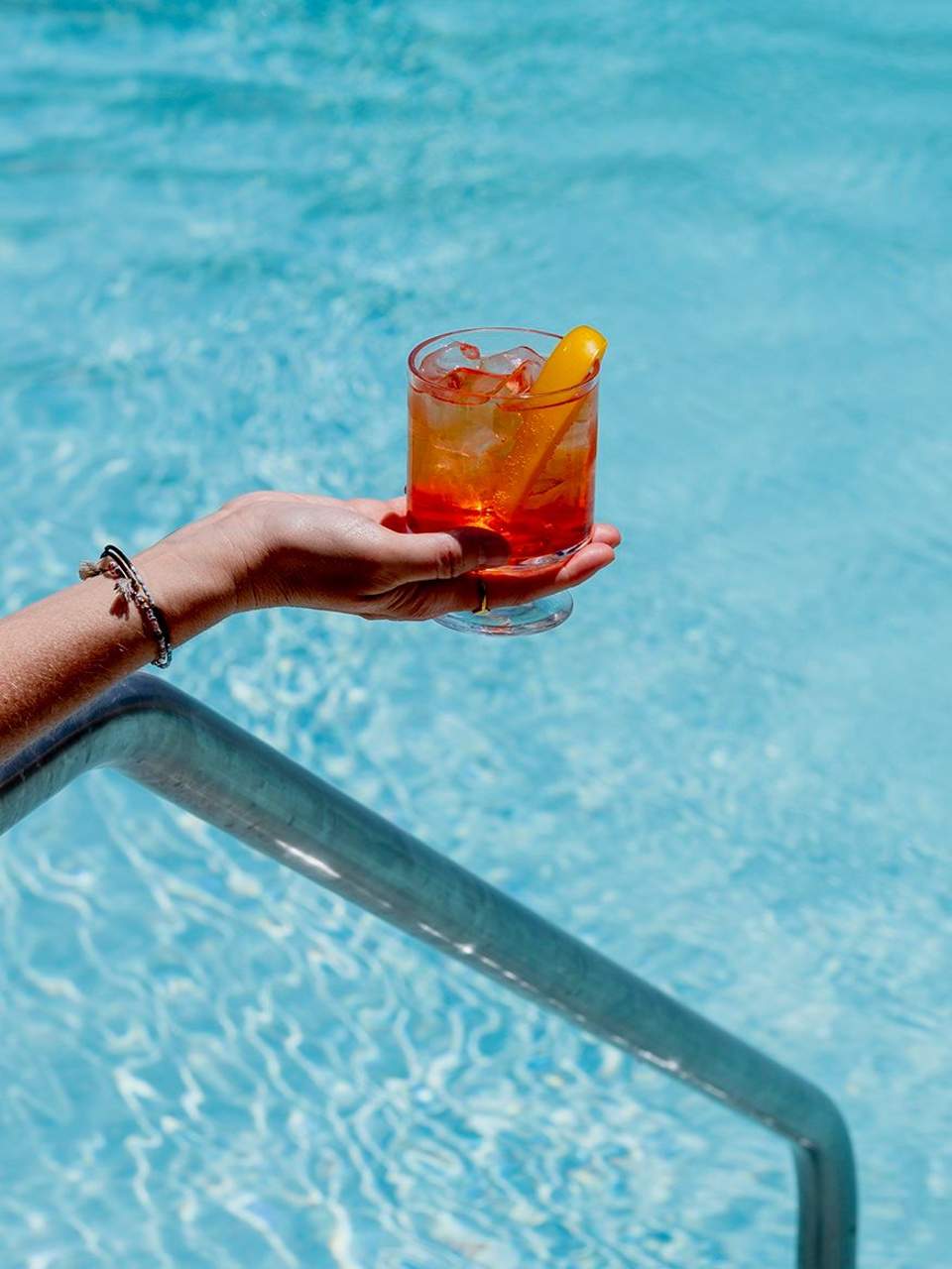 Hand holding a red cocktail with ice and lemon garnish over a swimming pool