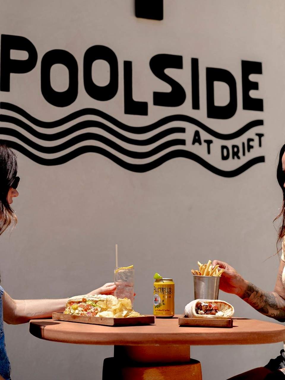 Two people sharing food at a round table with 'Poolside at Drift' signage behind them