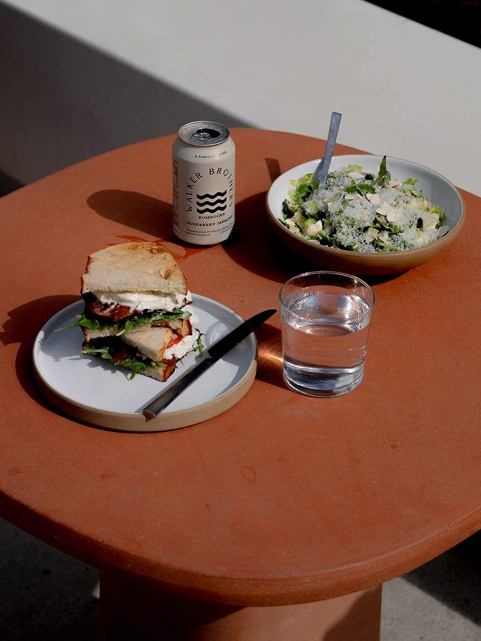 Outdoor dining table with sandwich, salad bowl, beverage can, and water glass on terracotta surface