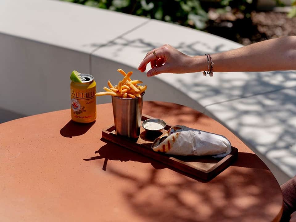 Hand reaching for food on terracotta table with sandwich, fries, and beverage visible