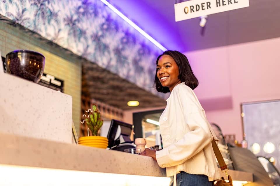 Smiling woman at modern cafe counter under purple neon lighting and ORDER HERE sign