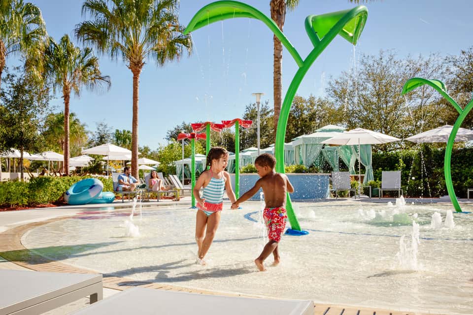 Children playing in shallow splash pad with green water features and palm trees at resort