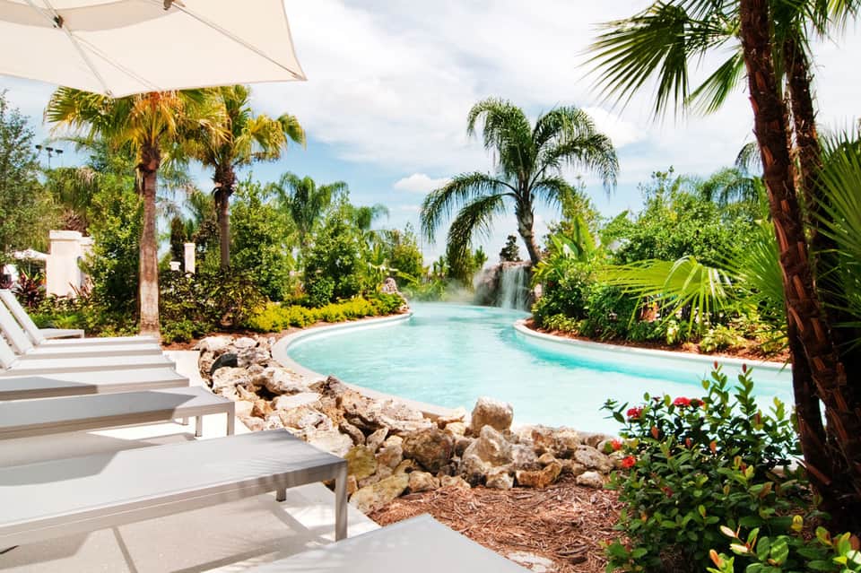 Resort lagoon-style pool with waterfall, palm trees, white umbrellas, and tropical landscaping