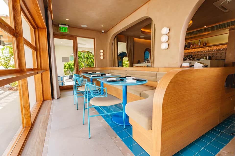 Modern restaurant interior with wooden curved booths, blue metal chairs, and large windows overlooking outdoor patio
