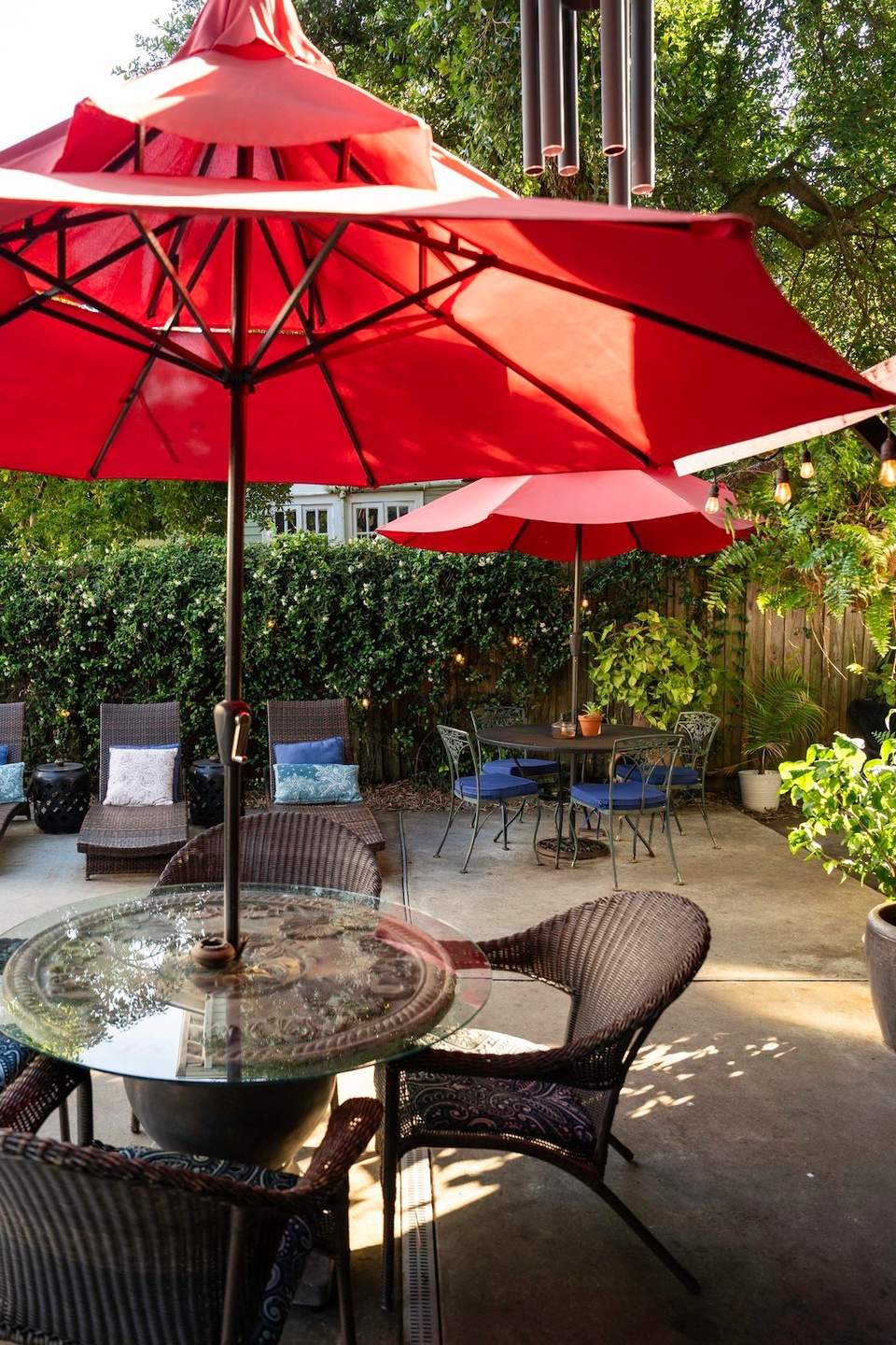 Outdoor patio with large red umbrellas, wicker chairs, and glass-top tables in a garden setting