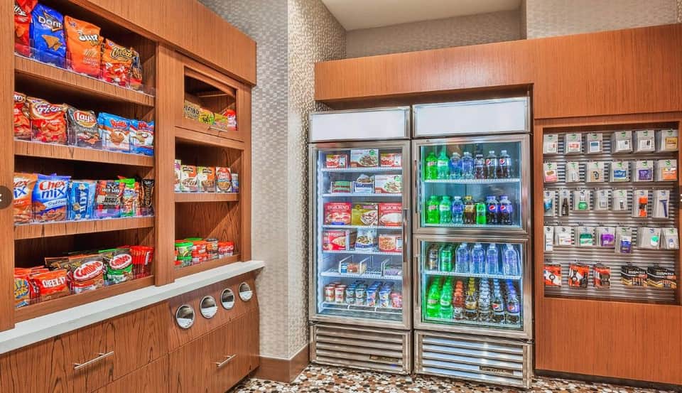 Hotel snack bar with wooden shelving displaying chips and beverages, refrigerated vending unit with drinks and packaged items