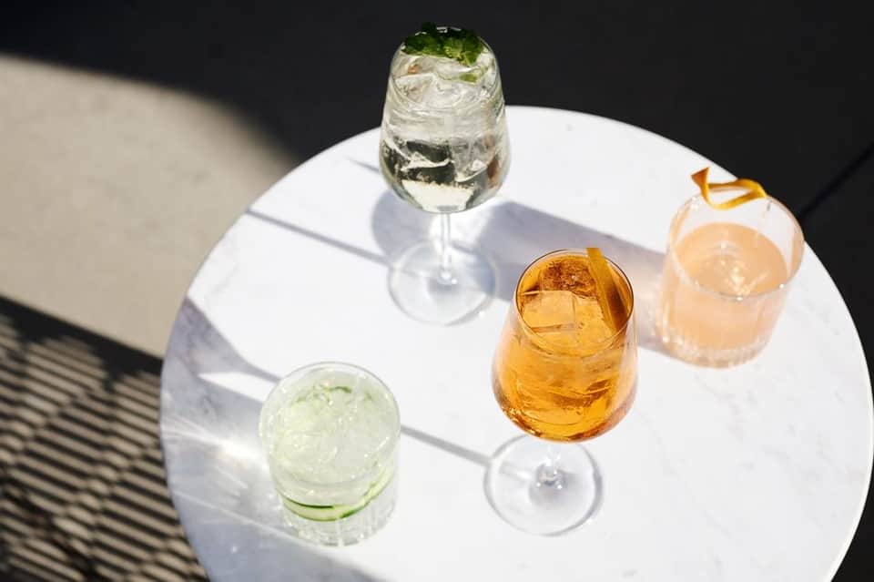 Cocktails on a white table including mojito, whiskey drink, and sparkling water in sunlight