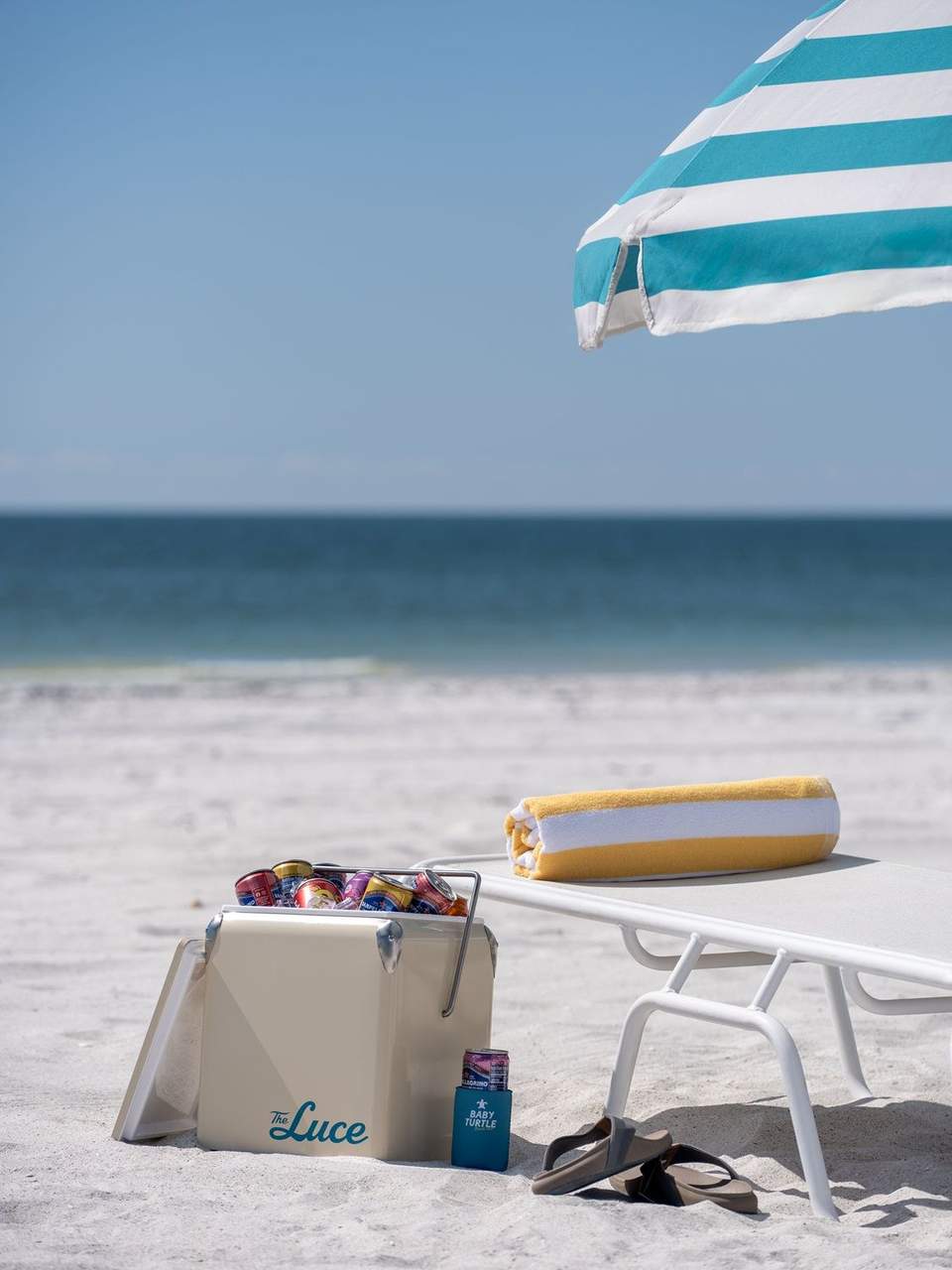 Beach lounge chair with striped umbrella, cooler with beverages, and rolled towel on sand