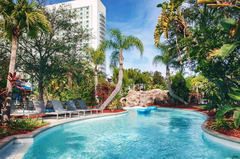 Tropical resort pool with turquoise water, palm trees, lounge chairs, and high-rise hotel building in background