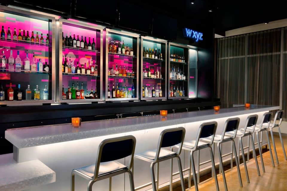 Contemporary bar with vibrant pink-lit shelving, spirits collection, white counter seating, and neon signage