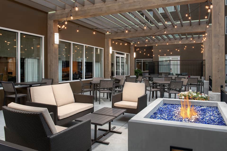 Outdoor patio with seating, fire pit with blue glass, string lights, wooden pergola, and glass doors to interior.
