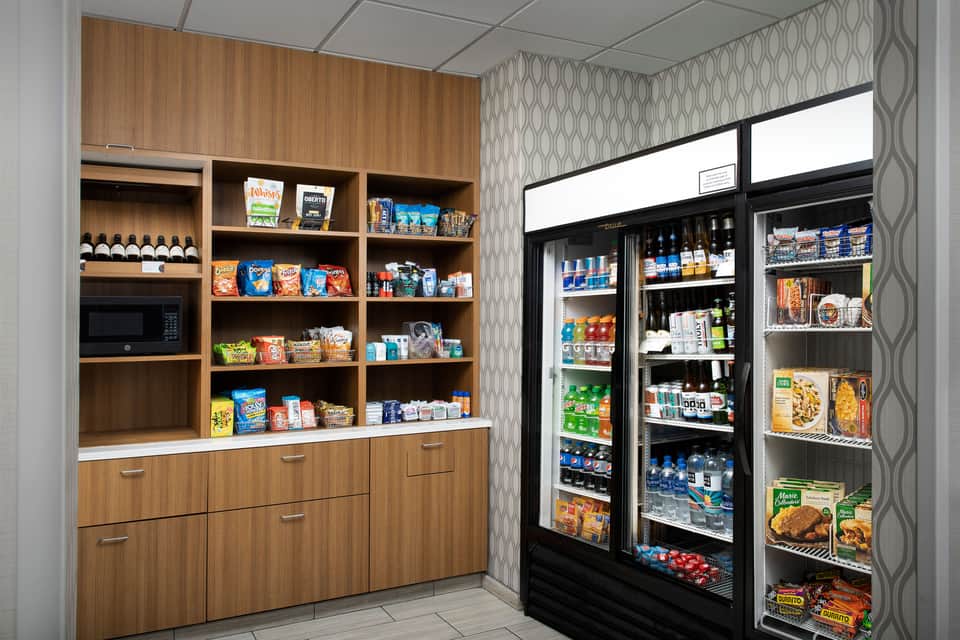 Hotel market area with wooden shelving, microwave, snacks, and refrigerated beverage coolers stocked with drinks and food.