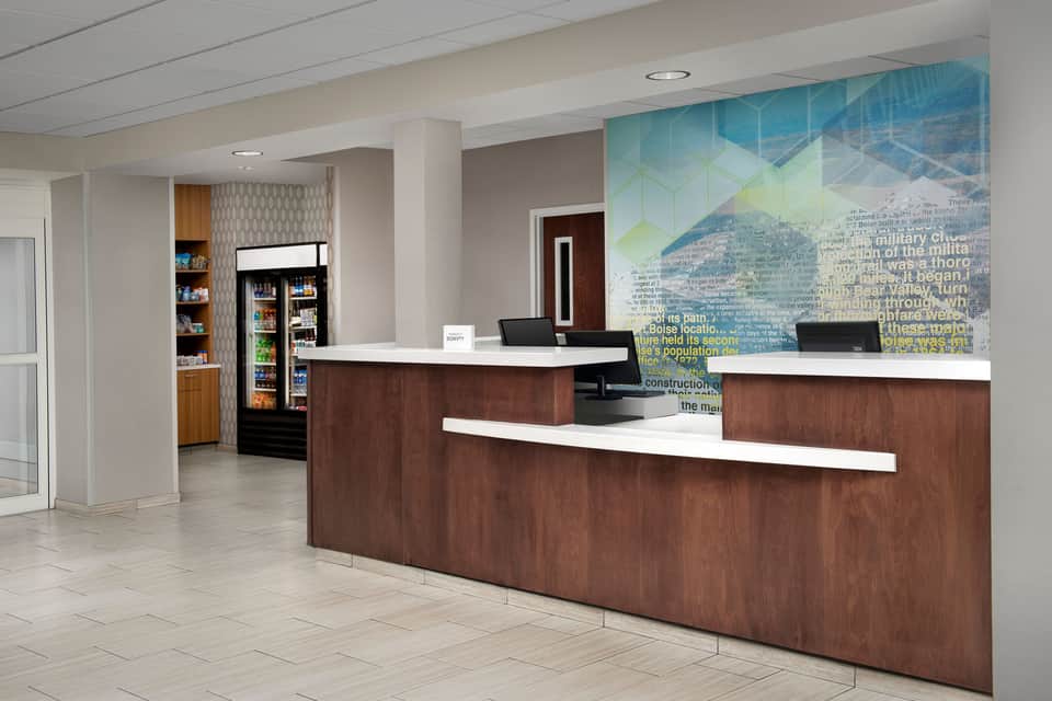 Modern hotel lobby with dark wood reception desk, beverage coolers, and informational wall mural