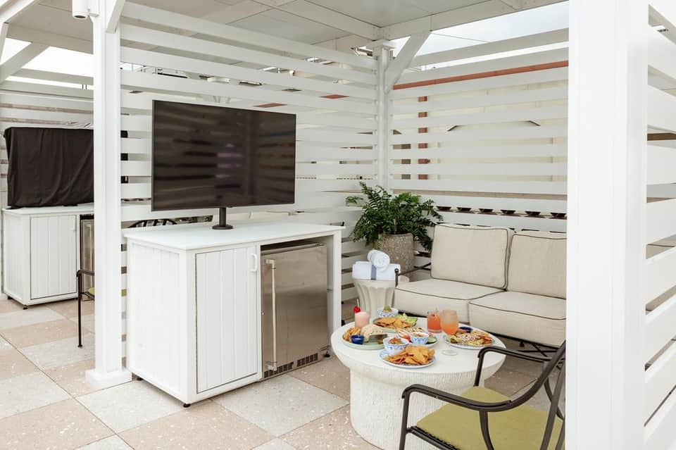 Covered outdoor lounge area with TV, mini-fridge, white furniture, and striped accent walls
