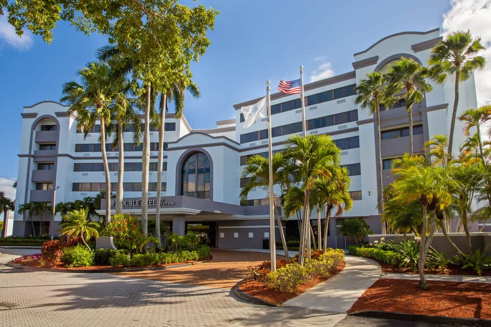 White multi-story hotel with curved architecture, palm trees, and American flag at entrance