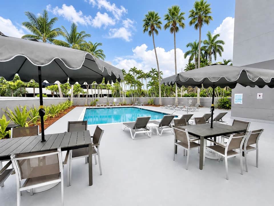 Resort pool area with loungers, black umbrellas, and tall palm trees under blue sky