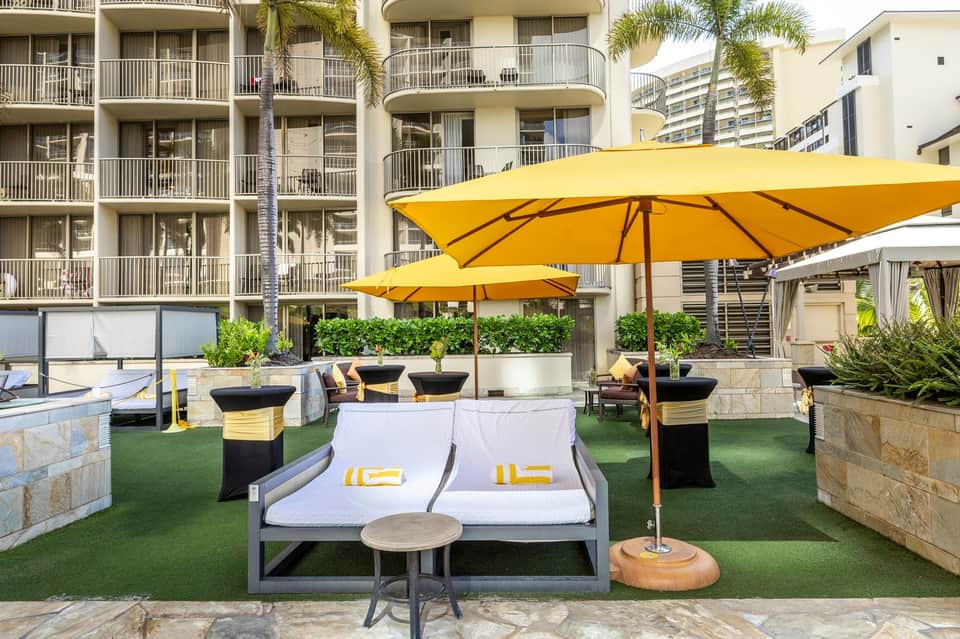 Outdoor lounge with yellow umbrellas, gray daybed, green turf, and multi-story hotel facade