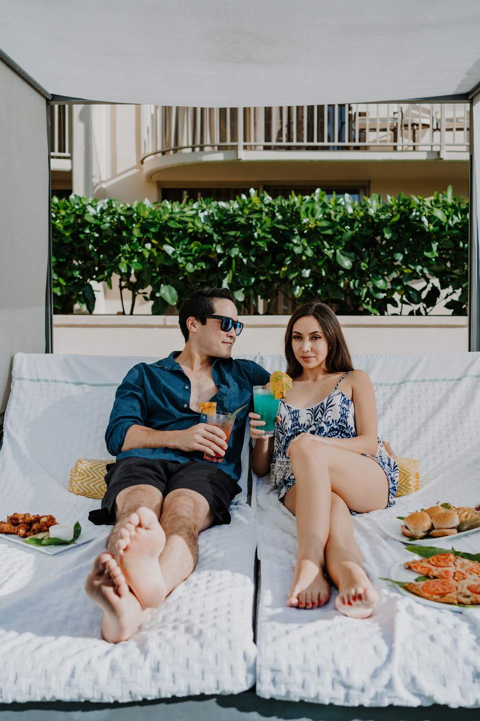 Couple relaxing on white daybed with drinks and food, tropical plants and balconies visible