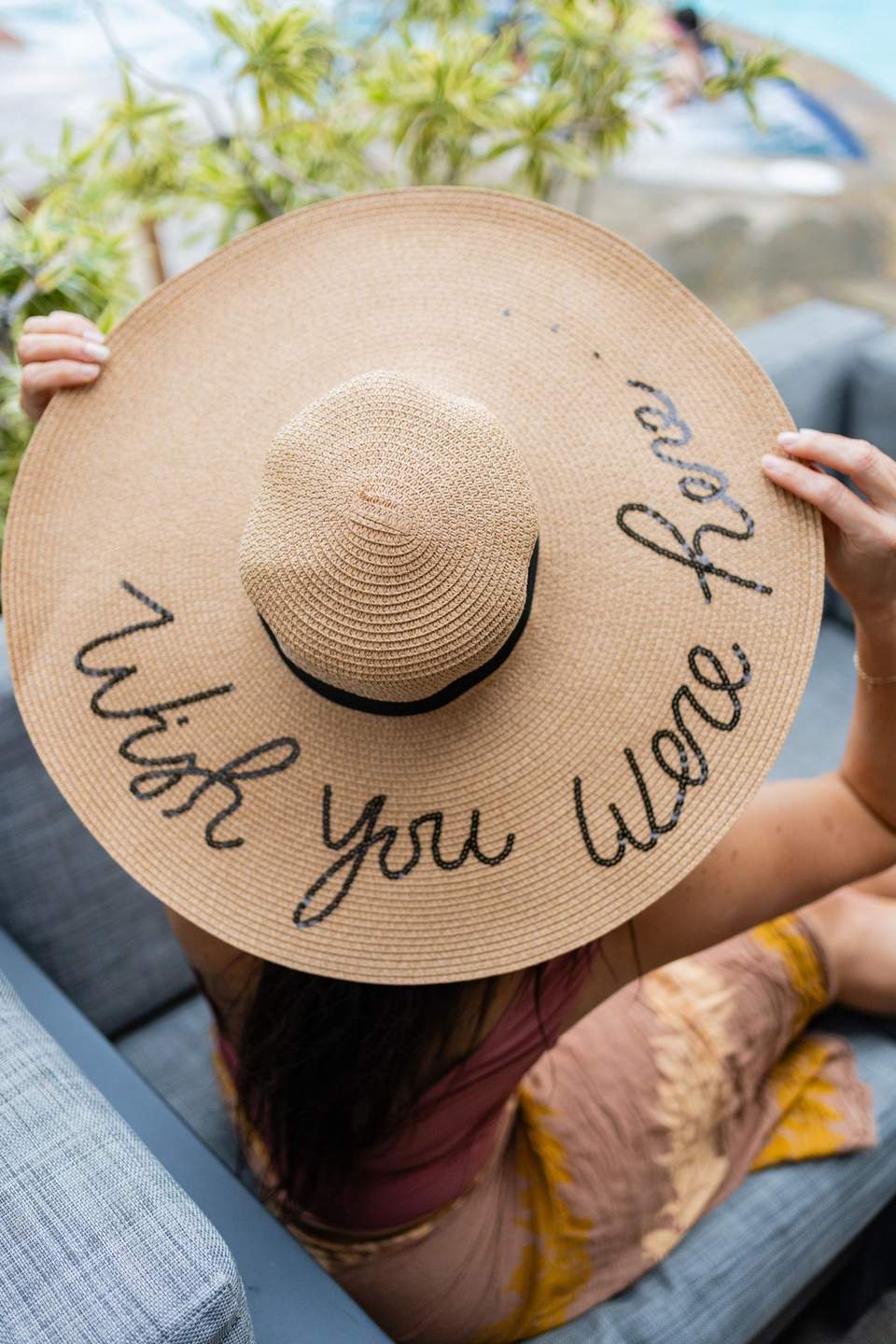 Person holding wide-brimmed straw hat with handwritten message 'Wish you were here'
