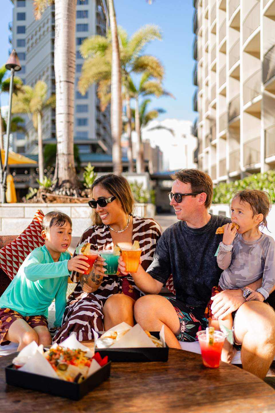 Family of four enjoying tropical drinks and food on resort terrace with palm trees