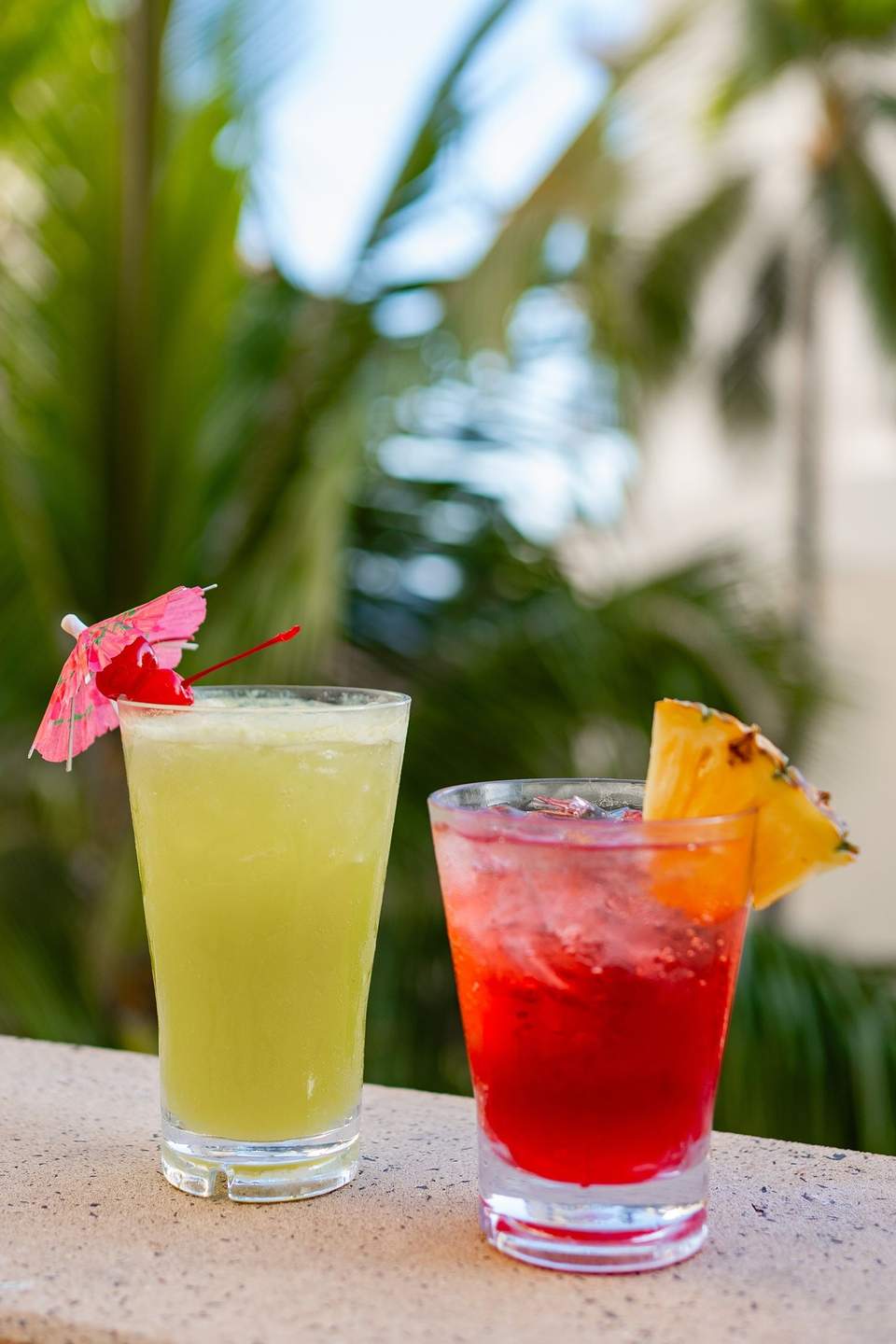 Two colorful tropical cocktails with umbrella and pineapple garnishes poolside