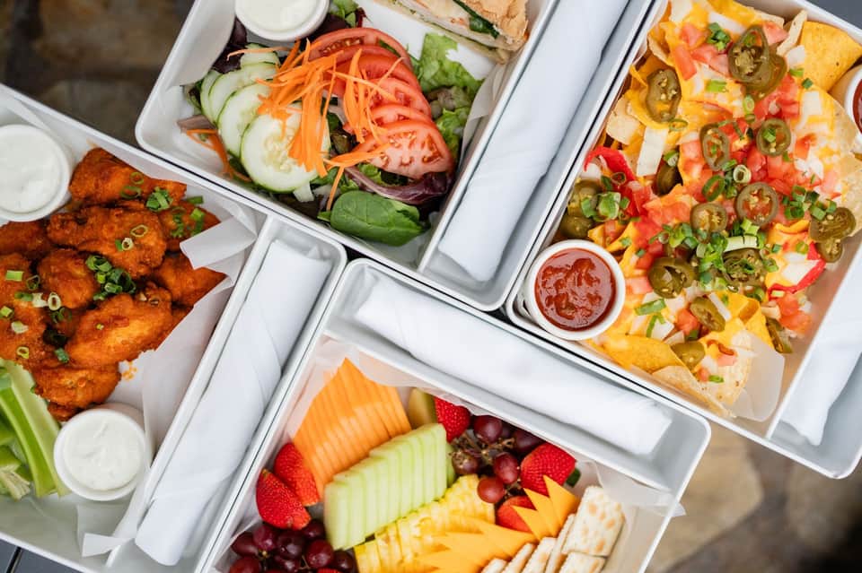Overhead view of catered food platters including wings, salad, nachos, fruit, and dips in white containers